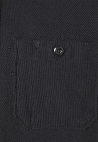 Tessuto lavorato a maglia nero con una superficie testurizzata e una tasca applicata con dettaglio a bottone. Il design è semplice e funzionale.
