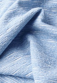 Hellblauer Stoff mit einem strukturierten Muster, das weiche, leicht erhabene Streifen und unregelmäßige weiße Akzente im gesamten Material aufweist.