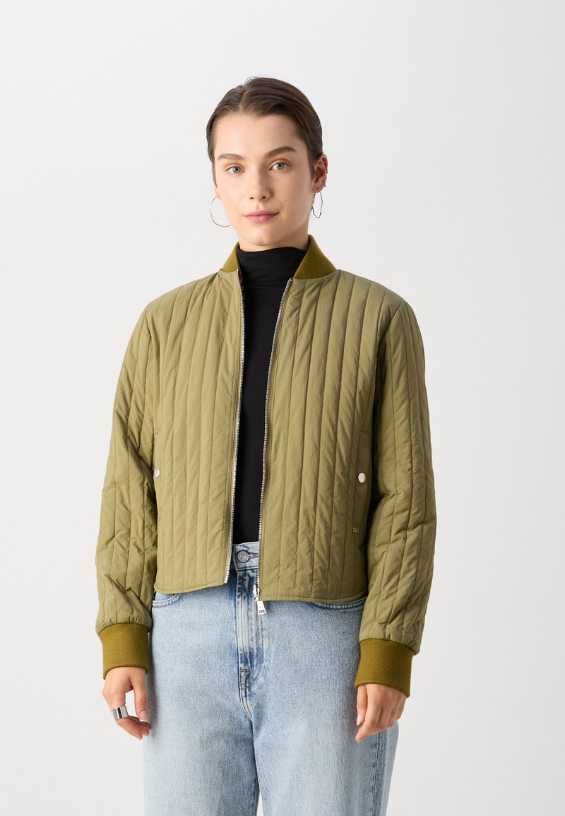 Veste bomber matelassée vert olive avec des poignets et un col côtelés, dotée d'une fermeture éclair à l'avant et de boutons-pression, associée à un jean bleu clair.