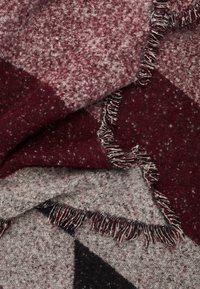 Couverture tissée avec un motif géométrique dans des tons bordeaux et gris, avec des bords à franges et un tissu texturé et doux.