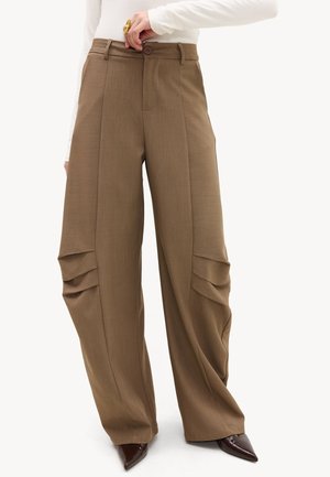 Pantalon marron à jambes larges avec détails plissés au niveau des genoux, bouton et fermeture éclair à l'avant, porté avec des chaussures marron pointues et un haut blanc à manches longues.