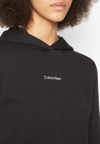 Persona che indossa una felpa con cappuccio nera con "Calvin Klein" stampato in piccolo testo bianco sul petto.