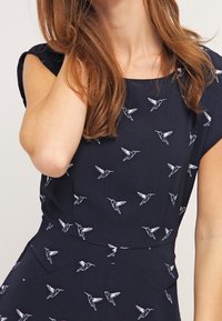 Marindress med mönster av kolibrier i vitt och rosa. Korta ärmar och en figurnära silhuett med subtil textur.