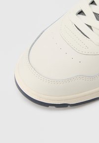 Κοντινό πλάνο στην μύτη ενός λευκού αθλητικού παπουτσιού με ανοιχτό γκρι ραφές, μικρές οπές διάτρησης, λευκά κορδόνια και γκρι χείλος σόλας.