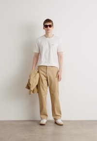 Cămașă albă cu mânecă scurtă, pantaloni largi bej, ținând în mână o jachetă bronz, purtând ochelari de soare maro și adidași albi, pe un fundal neutru.