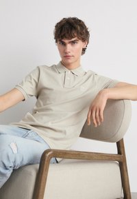 Μπεζ πόλο μπλούζα με κοντά μανίκια, τρεις κουμπότρυπες, υφή υφάσματος και μικρή λογότυπο. Φοριέται με φθαρμένα ανοιχτού μπλε τζιν.