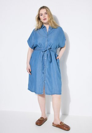 Denimhemdenkleid in Hellblau, mit kurzen Ärmeln, Knopfleiste, Bindegürtel und Seitentaschen. Getragen mit braunen Slippern.