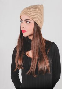 Berretto a cuffia beige a coste con punta, indossato da una persona. I capelli sono lunghi e castani, abbinati a un maglione nero a coste a maniche lunghe.