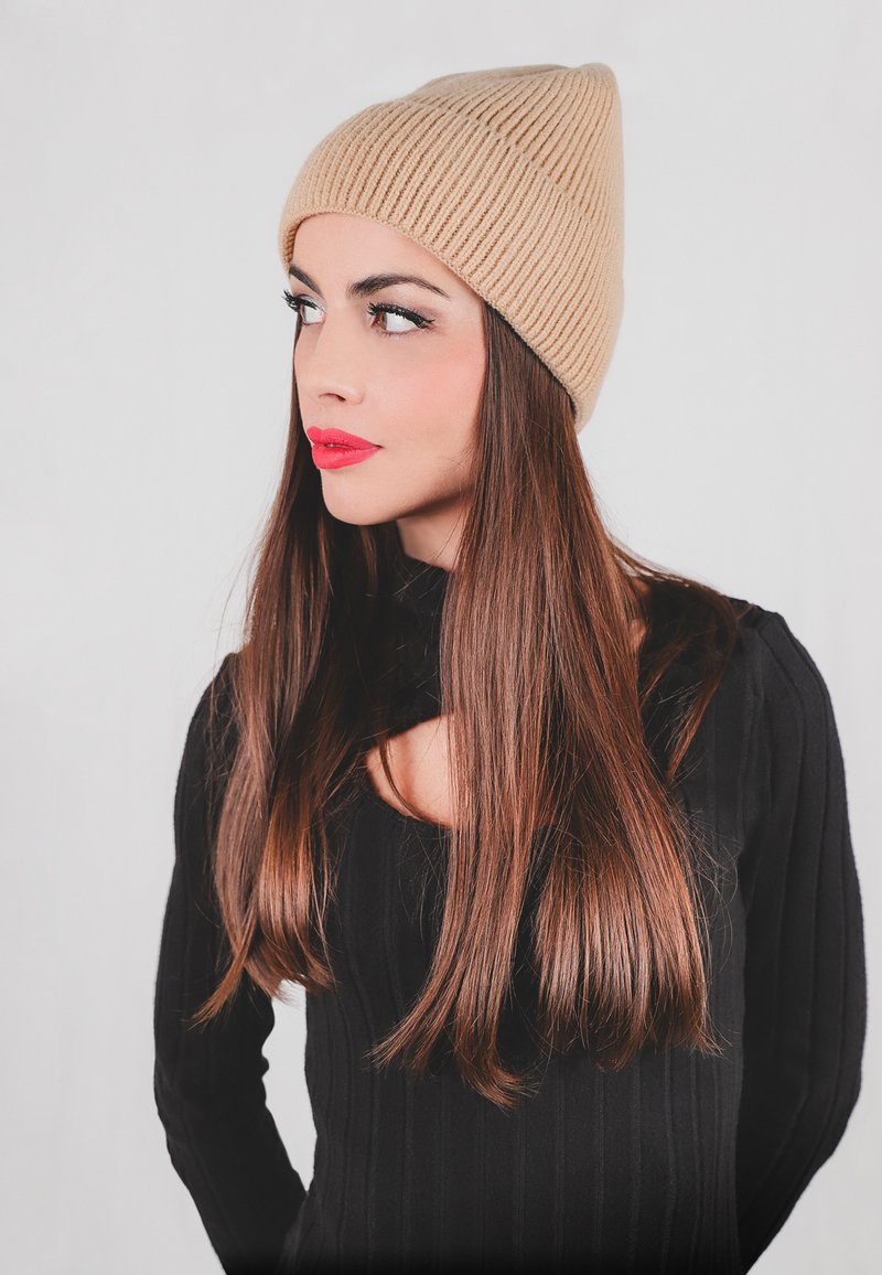Berretto a cuffia beige a coste con punta, indossato da una persona. I capelli sono lunghi e castani, abbinati a un maglione nero a coste a maniche lunghe.