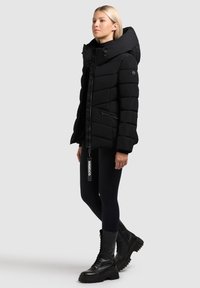 Jachetă puf black cu o glugă mare, închidere cu fermoar și design quilted. Dispune de buzunare laterale și un etichetă de marcă care atârnă de fermoar.