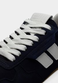 Σουέντ ναυτικό αθλητικό παπούτσι με λευκά κορδόνια και δύο λευκές πλευρικές ρίγες. Διαθέτει υφή στη γλώσσα και λαστιχένια σόλα. Κοντινή άποψη του πάνω μέρους.