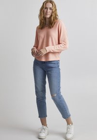 Licht zalmkleurig gebreid sweater met lange mouwen, gecombineerd met gescheurde blauwe jeans en witte sneakers, tegen een neutrale achtergrond.