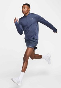 Marineblå langærmet atletisk trøje med en halv lynlås og tommelhuller, parret med mørkegrå løbeshorts og hvide sneakers. Glat stofstruktur.