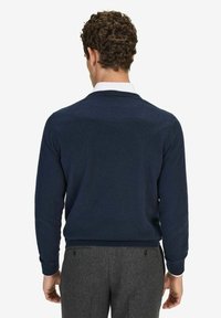 Marinblå tröja med rundad halsringning, ribbade muddar och nederkant, tillverkad av ett strukturerat stickmaterial. Bärs över en vit skjorta med krage.