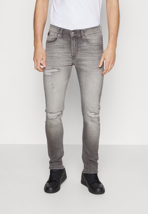 Homme portant un jean slim gris délavé avec des patchs usés, des baskets noires et un t-shirt blanc, debout sur un fond uni.