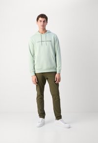 Felpa verde chiaro con la scritta 'COPENHAGEN' stampata in grigio, grande tasca frontale, abbinata a pantaloni verde oliva e sneakers bianche.