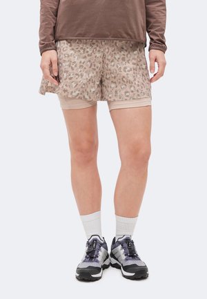 Underdel af person iført beige mønstret nederdel-shorts, brun langærmet top, hvide sokker og grå-sorte sportssko, stående på lys baggrund.