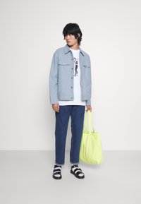 Chaqueta de mezclilla azul claro, camiseta gráfica blanca, jeans oscuros, sandalias en blanco y negro, sosteniendo una tote bag amarilla brillante. Texturas suaves, ajuste casual.