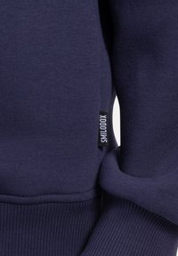 Sudadera de canalé azul marino con mangas y puños, y una pequeña etiqueta de tela negra que dice "SMILODOX" cosida en la costura lateral.