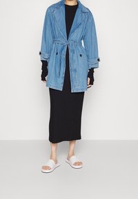 Riflový trenčkot s páskem v pase, velkými klopami a knoflíkovými detaily, vrstvený přes černé šaty a doplněný bílými pantoflemi.