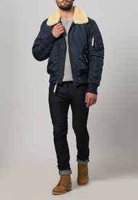 Veste bomber en nylon marine avec un col en fausse fourrure crème, poches zippées, poignets côtelés et une coupe décontractée, associée à un jean foncé et des bottes beiges.