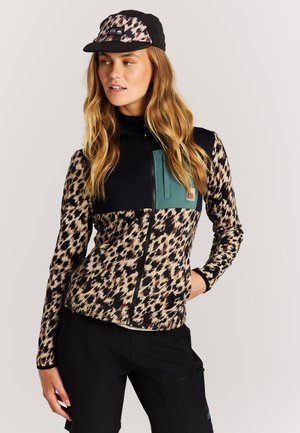 Vrouw met een jaguarprint ritsjack met zwarte en turquoise accenten, een bijpassende pet en zwarte broek, kijkt opzij met haar hand in de zak.