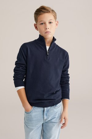 Tamsiai mėlynas džemperis su užtrauktuku, pagamintas iš minkštos medžiagos, su ilgomis rankovėmis ir austo audinio apvadais rankogaliuose. Dėvimas ant baltos marškinės su šviesiai mėlynomis džinsais.