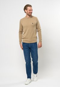 Beige gebreide trui met lange mouwen, ronde halslijn en kleine logo-accent. Gecombineerd met blauwe denim jeans en witte sneakers.