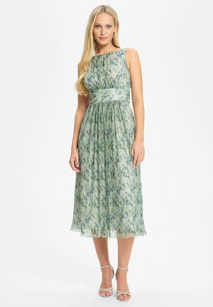 Femme aux longs cheveux blonds portant une robe midi verte à fleurs sans manches et des talons argentés à brides, debout devant un fond blanc.