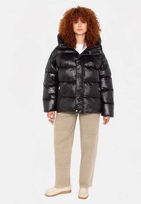 Giacca a vento nera con texture trapuntata lucida, cappuccio grande, zip frontale e chiusura a scatto. Abbinata a pantaloni beige chiaro e scarpe bianco sporco.