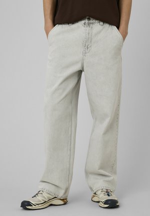 Personne portant un jean ample gris clair avec les mains dans les poches et des baskets blanches sportives avec des touches noires et beiges.