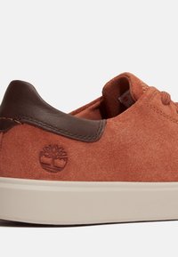 Zapatillas de ante marrón con una pestaña de talón de cuero marrón oscuro, suela beige y logotipo de árbol en relieve en el lateral.