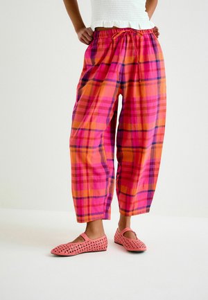 Person wearing loose pink and orange plaid pants with drawstring waist and pink perforated slip-on shoes, standing with hands on hips.