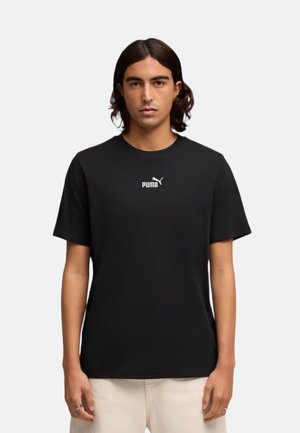 Joven con cabello oscuro hasta los hombros, vistiendo una camiseta negra de Puma y pantalones claros frente a un fondo llano.