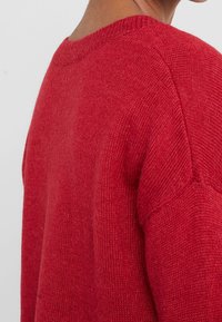 Pull en laine rouge avec un col rond et des détails côtelés au niveau du col. Texture douce et coupe décontractée. Présenté sous un angle de côté et de dos.