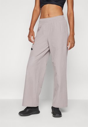 Vrouw die lichtgrijze wijde broek met een elastische tailleband en zwarte sneakers draagt, staand tegen een effen lichte achtergrond.