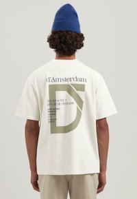 Camiseta blanca de manga corta con estampado gráfico verde en la parte posterior, que presenta texto y un diseño estilizado de la "D", fabricada en algodón texturizado.