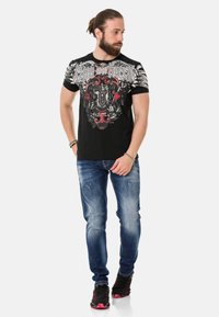 Schwarzes T-Shirt mit einem detaillierten grafischen Design in Weiß, Rot und Grau; kombiniert mit hellblauen Jeans und schwarzen Turnschuhen mit roten Akzenten.