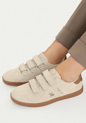 Lyse beige casual sneakers med tre velcroremme og brune såler, båret med opkaldte brune bukser.