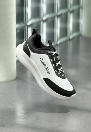 Biało-czarny sportowy but z logo Calvin Klein, białymi sznurówkami i grubą podeszwą, unoszący się nad szarą podłogą w pobliżu szklanych bloków.