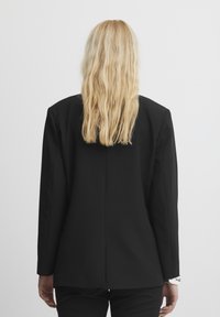 Zwarte op maat gemaakte blazer met een notch-kraag, gestructureerde schouders en een enkele split aan de achterkant. Gladde stof met een matte afwerking.