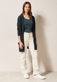 Mörkgrön lång cardigan, åtsittande mörk topp, krämfärgade byxor och vita sneakers; mjuk textur och fickor på cardigan.