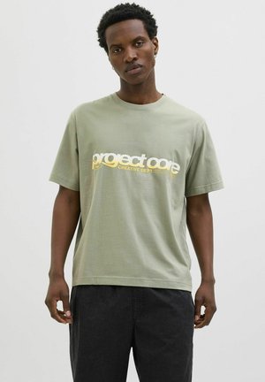T-shirt en coton vert olive à manches courtes, comportant un texte en gras blanc et jaune "project core CREATIVE DEPT." sur le devant.