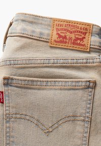 Des jeans en denim bleu clair présentent une étiquette en cuir avec des accents graphiques rouges. Deux poches arrière avec des coutures jaunes et le logo Levi's visible.