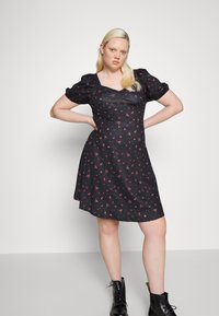 Vestido preto com pequenos padrões florais vermelhos, mangas curtas em balão, corpete justo e saia rodada; usado com botas pretas de atacador.