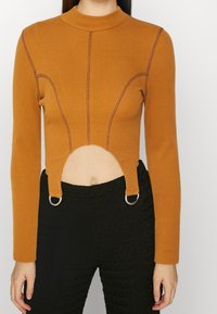 Rost-orange långärmad topp med ribbad textur, med en urklippt sektion och metalringar för knäppning. Förstärkningsdetaljer i sömmarna.