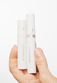 Deux contenants cosmétiques cylindriques blancs avec des accents argentés et un logo "W" en relief, tenus dans une main. L'un est légèrement plus grand que l'autre.