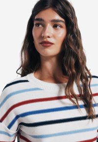 Gestreifter Strickpullover in Weiß, mit horizontalen Streifen in Rot, Blau und Marineblau. Klassischer Rundhalsausschnitt und gerippte Struktur.