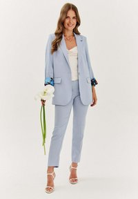 Světle modrý oblek na míru s formálním sakem a slim fit kalhoty. Bílé tričko pod ním, květinové akcenty na rukávech, drží květinu.