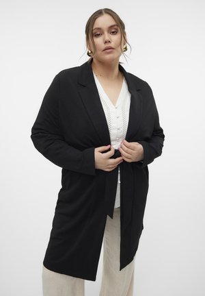 Black long-sleeve blazer with a belted waist, tailored design, and lapel collar. Worn over a white button-up shirt, paired with beige trousers.